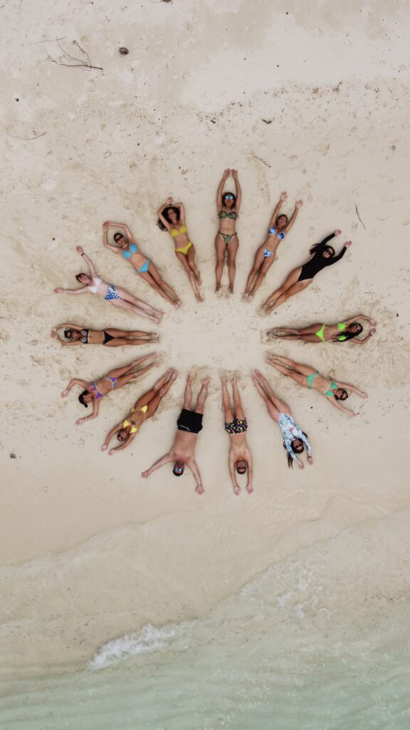 Grupo de personas acostadas en la arena formando un círculo con sus cuerpos en una playa tropical.