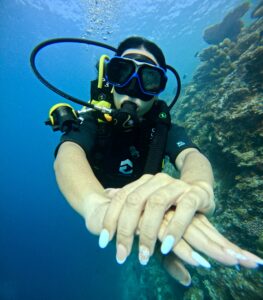 Mujer buceando con equipo de oxígeno bajo el agua, extendiendo sus manos con uñas decoradas mientras explora un arrecife de coral.