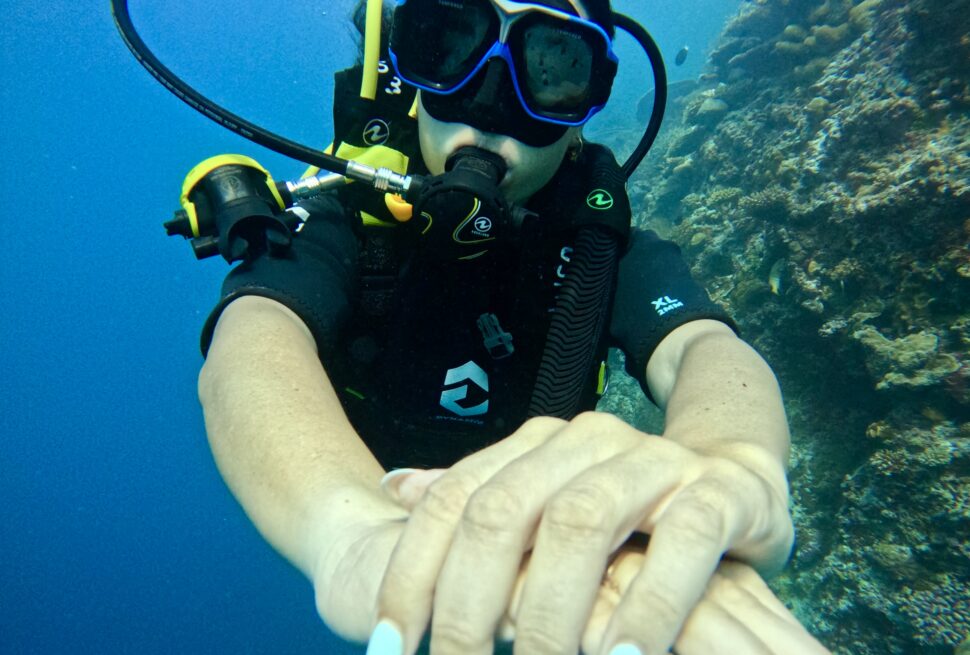 Mujer buceando con equipo de oxígeno bajo el agua, extendiendo sus manos con uñas decoradas mientras explora un arrecife de coral.