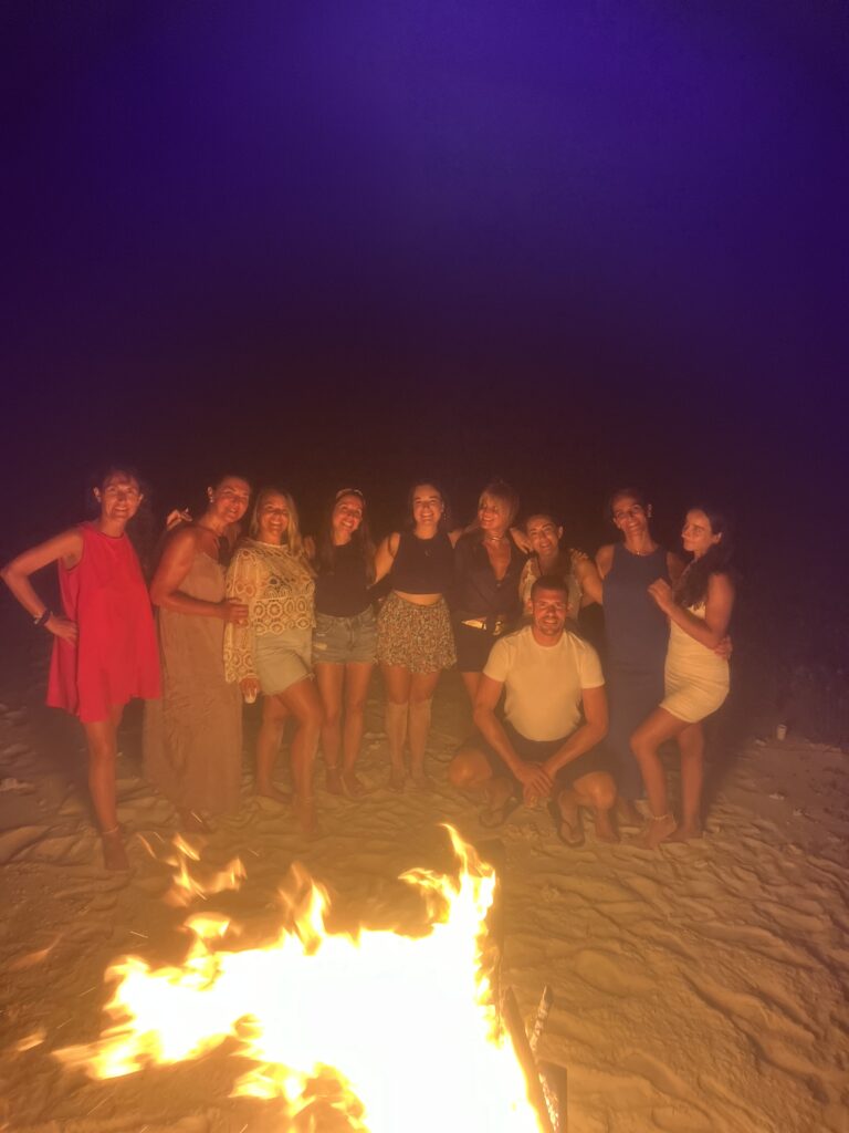 Grupo de personas reunidas alrededor de una fogata en la playa durante la noche.