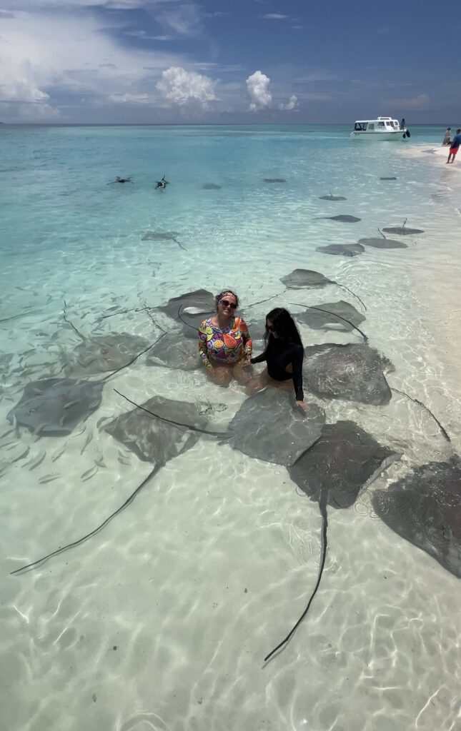 Dos mujeres sentadas en aguas cristalinas rodeadas de mantarrayas en una playa tropical.