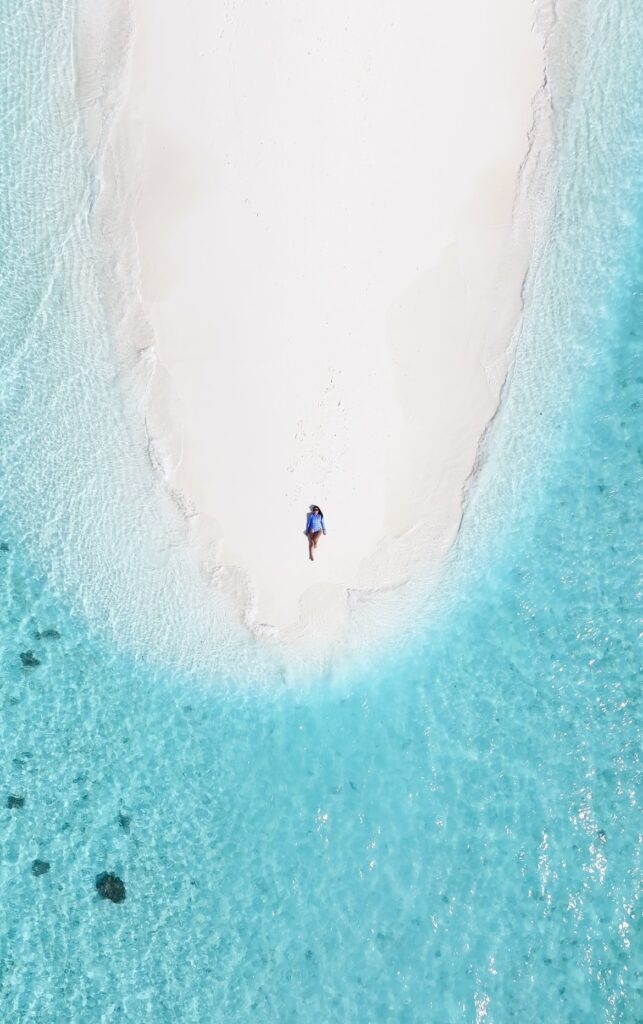 Mujer acostada en una pequeña isla de arena blanca rodeada de aguas cristalinas vistas desde un ángulo aéreo