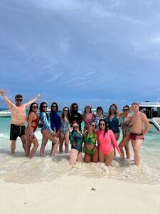 Grupo de personas posando en la orilla de una playa con el mar y varias embarcaciones de fondo.