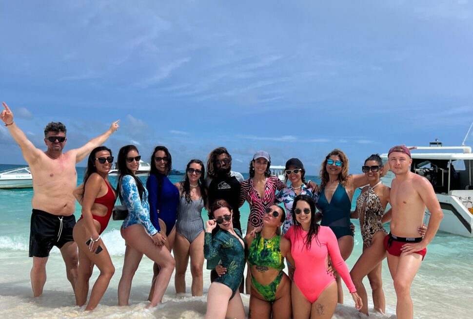 Grupo de personas posando en la orilla de una playa con el mar y varias embarcaciones de fondo.