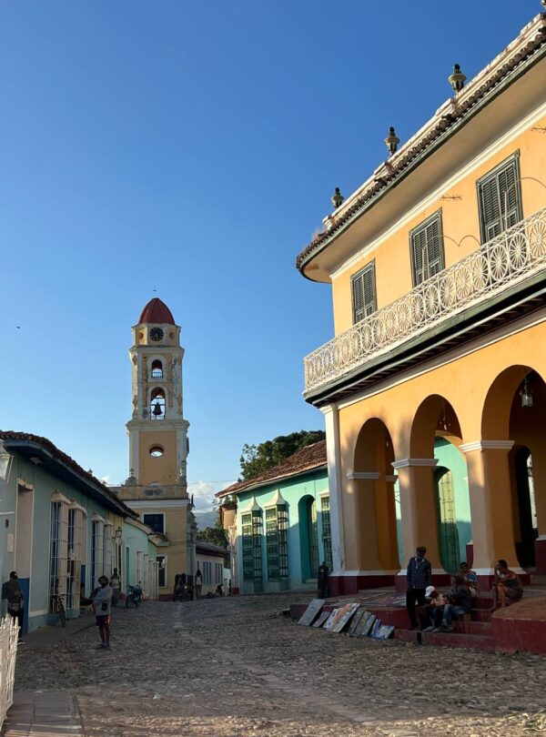 Vista de una plaza en Trinidad, Cuba, con una gran palmera en primer plano, una iglesia con torre y un edificio colonial amarillo con balcones.
