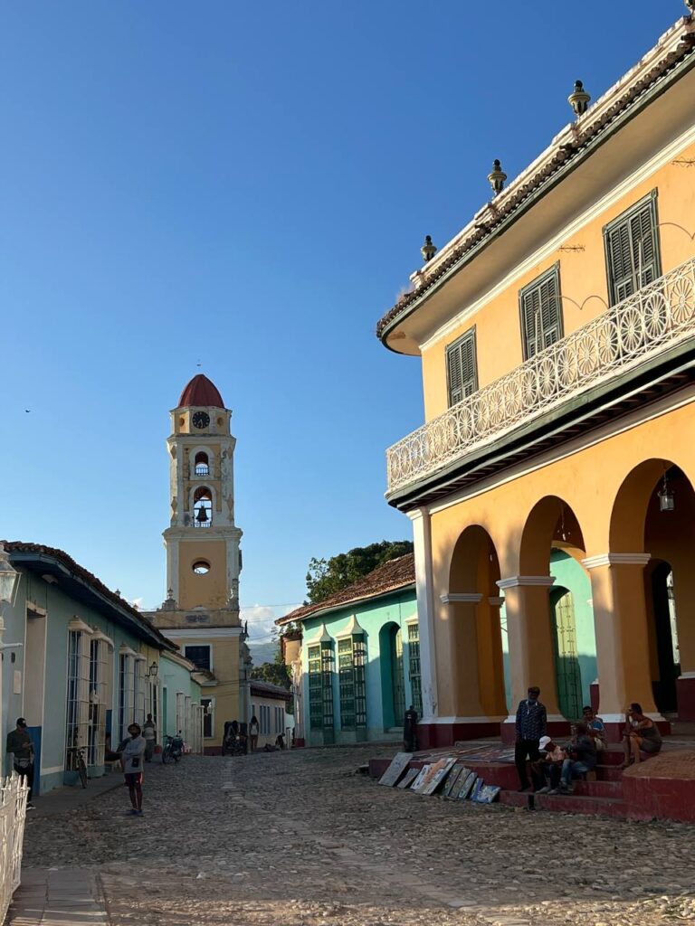 Vista de una plaza en Trinidad, Cuba, con una gran palmera en primer plano, una iglesia con torre y un edificio colonial amarillo con balcones.