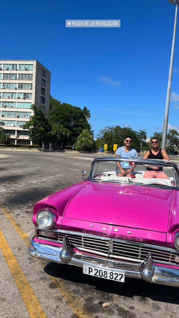 Auto clásico descapotable de color rosa con dos hombres sonrientes a bordo, estacionado en la Plaza de la Revolución en La Habana, Cuba.