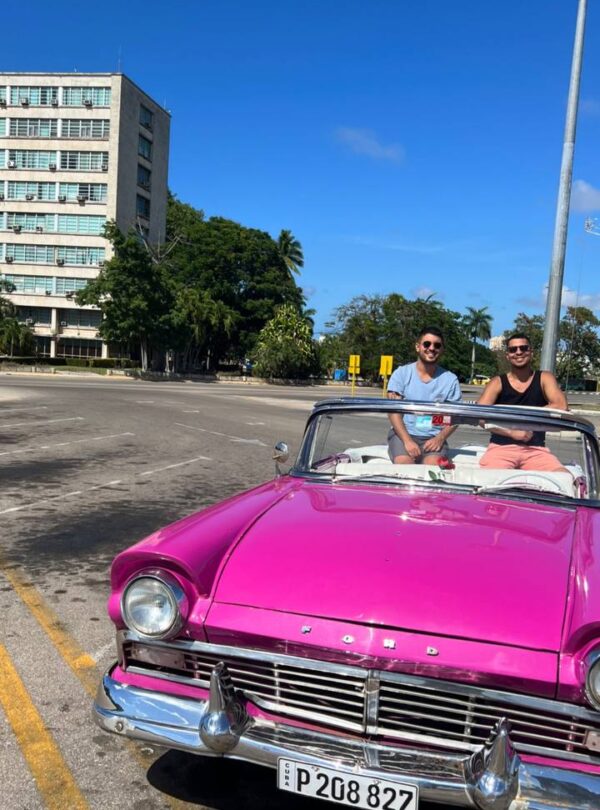 Auto clásico descapotable de color rosa con dos hombres sonrientes a bordo, estacionado en la Plaza de la Revolución en La Habana, Cuba.