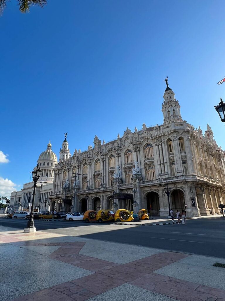 Gran Teatro de La Habana y el Capitolio bajo un cielo azul, con coco-taxis amarillos estacionados en la avenida.