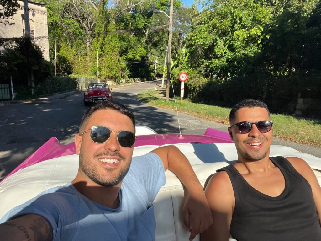 Dos hombres sonrientes con gafas de sol viajando en un auto clásico descapotable rosa por una calle rodeada de vegetación, con otro coche antiguo rojo en el fondo