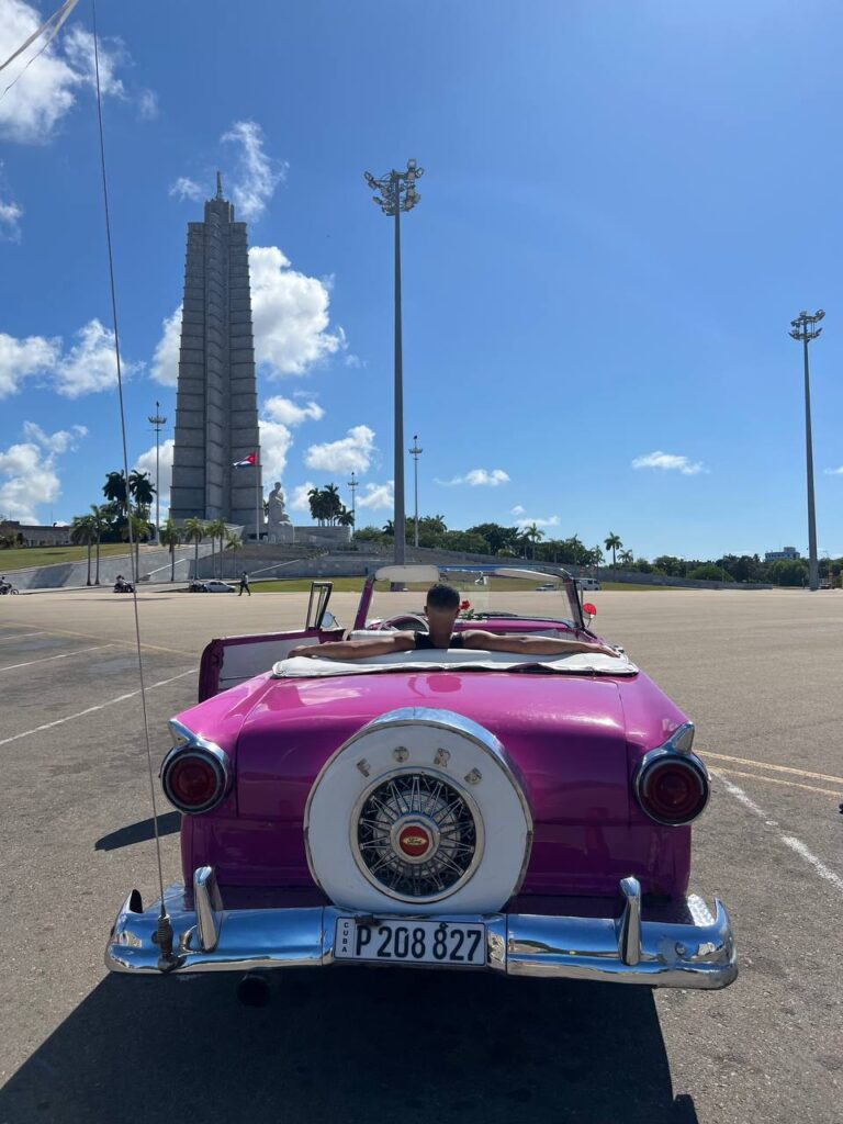 Auto clásico descapotable rosa con un pasajero relajado en su interior, estacionado frente al Monumento a José Martí en la Plaza de la Revolución, La Habana.