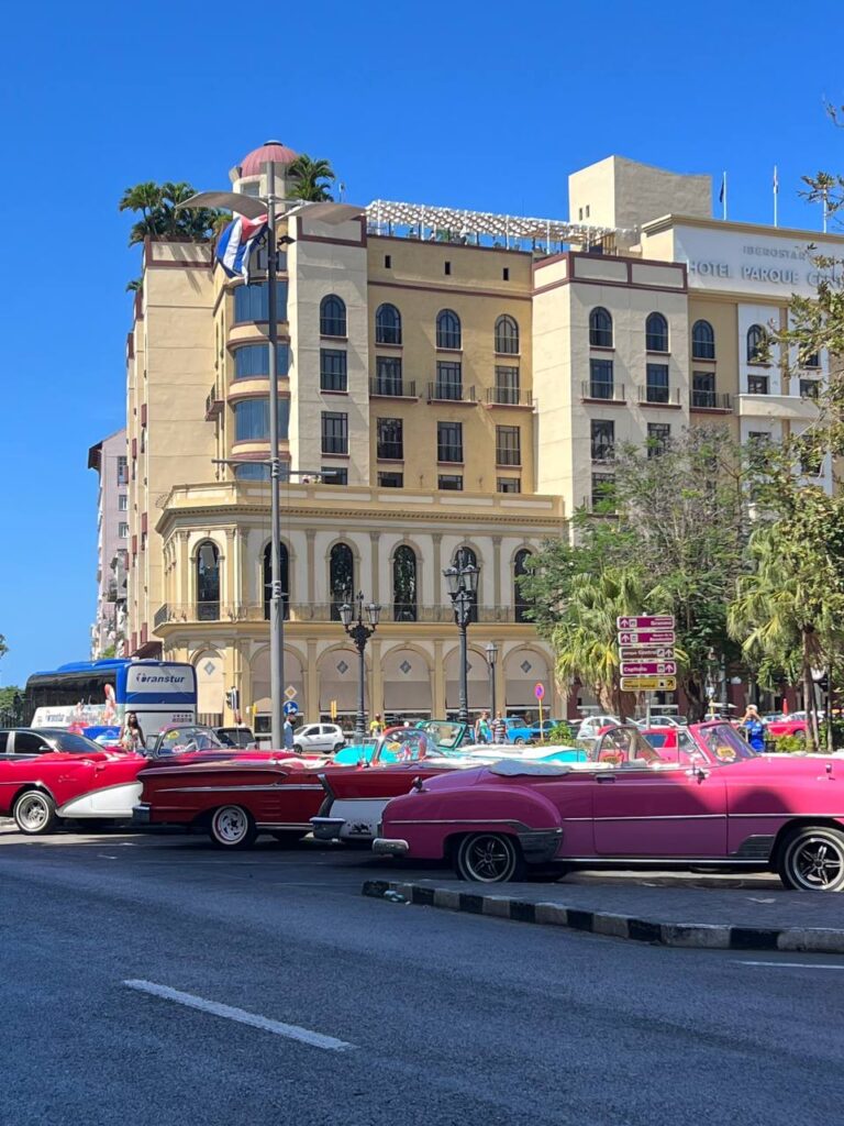 Autos clásicos descapotables de colores vibrantes estacionados en una calle de La Habana, con el Hotel Iberostar Parque Central de fondo.