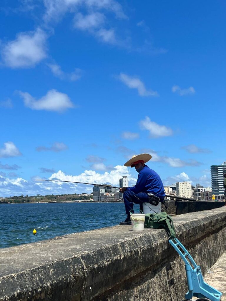 Hombre con sombrero de ala ancha pescando en el Malecón de La Habana bajo un cielo azul con nubes.