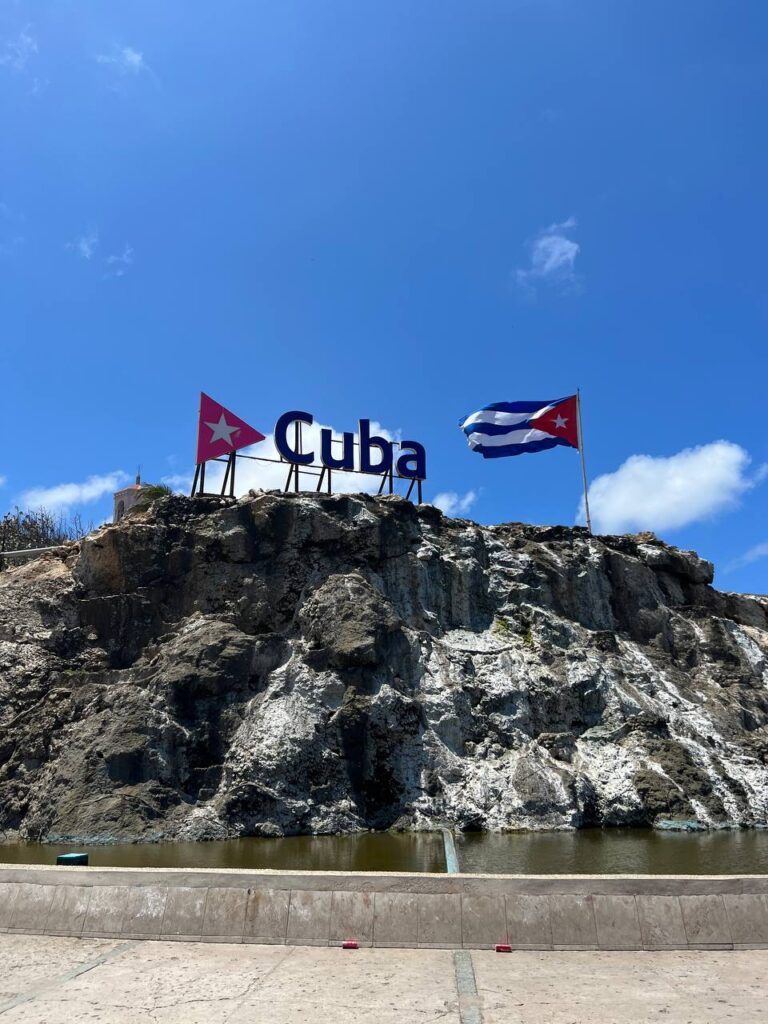 Cartel con la palabra "Cuba" y la bandera cubana ondeando sobre una colina rocosa bajo un cielo azul despejado