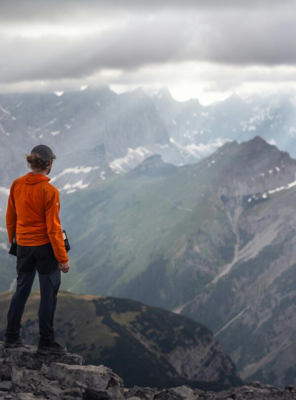 Persona con chaqueta naranja observando un paisaje montañoso rodeado de nubes grises y picos nevados