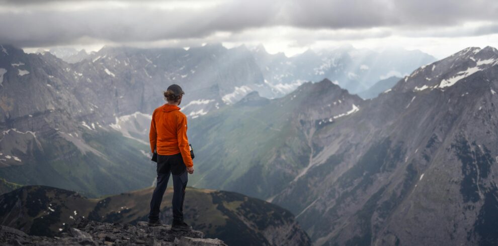 Persona con chaqueta naranja observando un paisaje montañoso rodeado de nubes grises y picos nevados