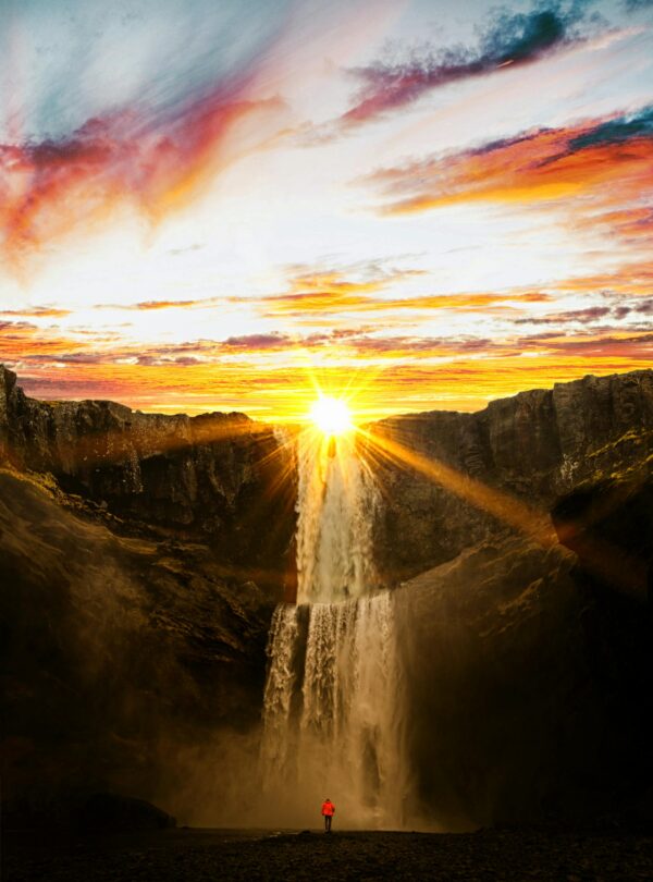 Persona con chaqueta roja frente a una imponente cascada iluminada por el sol durante un atardecer espectacular