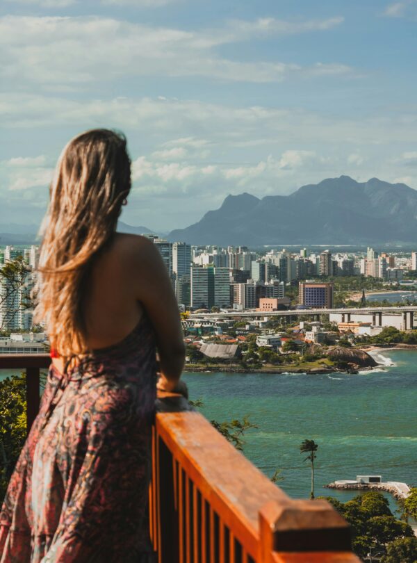 Mujer contemplando una ciudad costera con montañas al fondo desde un mirador de madera.