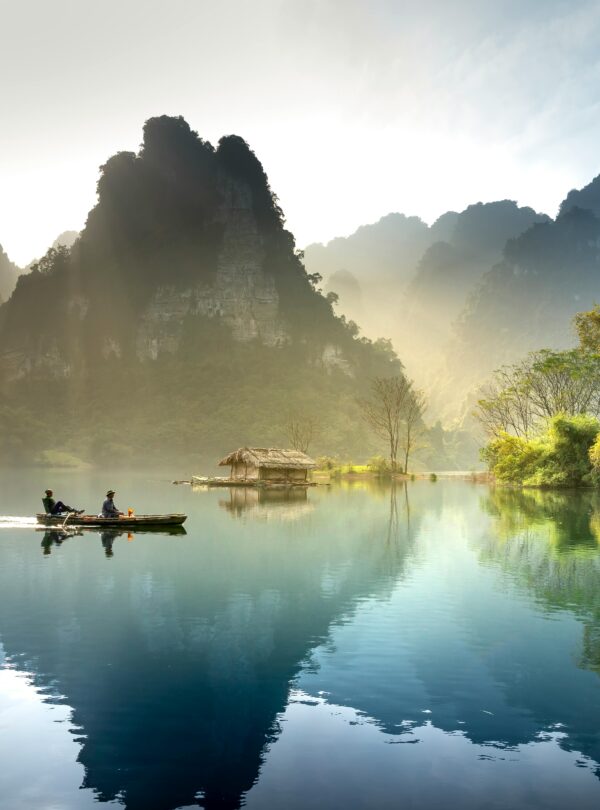 Dos personas en un bote navegando en un lago tranquilo, rodeado de majestuosos picos montañosos, vegetación exuberante y una cabaña en la orilla iluminada por la luz del amanecer