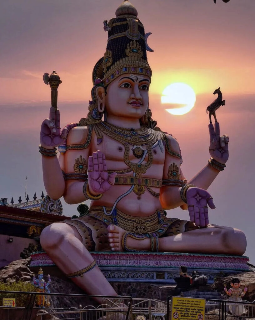 Estatua gigante del dios hindú Shiva con el sol poniéndose detrás de ella, en un entorno de templo.