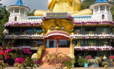 Estatua gigante dorada de Buda en el Templo Dorado de Dambulla, Sri Lanka, con un edificio decorado y torres azules a sus lados.