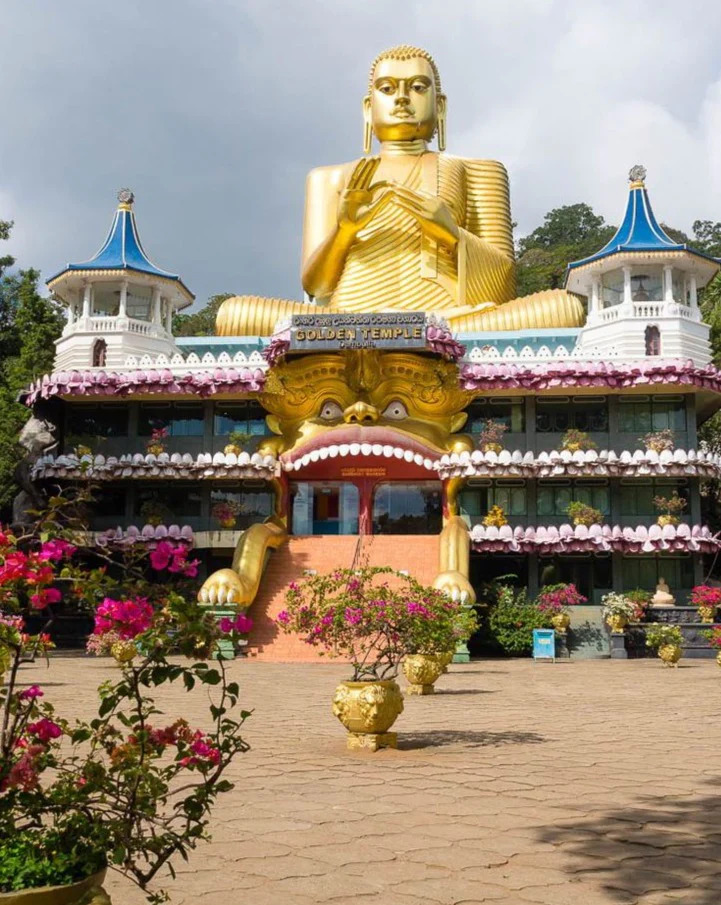 Estatua gigante dorada de Buda en el Templo Dorado de Dambulla, Sri Lanka, con un edificio decorado y torres azules a sus lados.