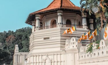Vista exterior del Templo del Diente en Kandy, Sri Lanka, con arquitectura blanca junto a un estanque y banderas budistas.