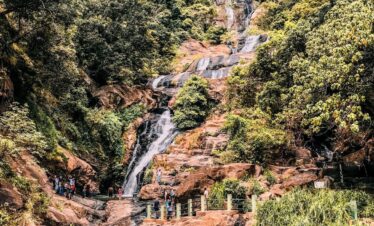 Cascada Ravana en Sri Lanka fluyendo entre rocas y vegetación densa, con visitantes caminando por senderos cercanos.