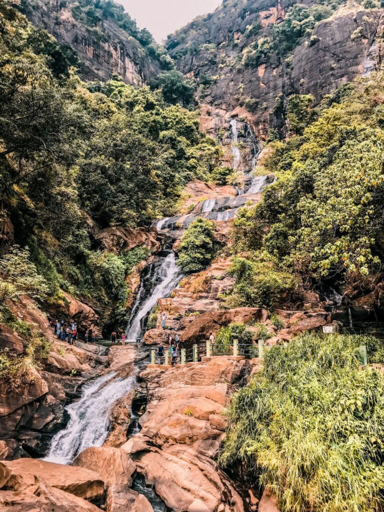 Cascada Ravana en Sri Lanka fluyendo entre rocas y vegetación densa, con visitantes caminando por senderos cercanos.