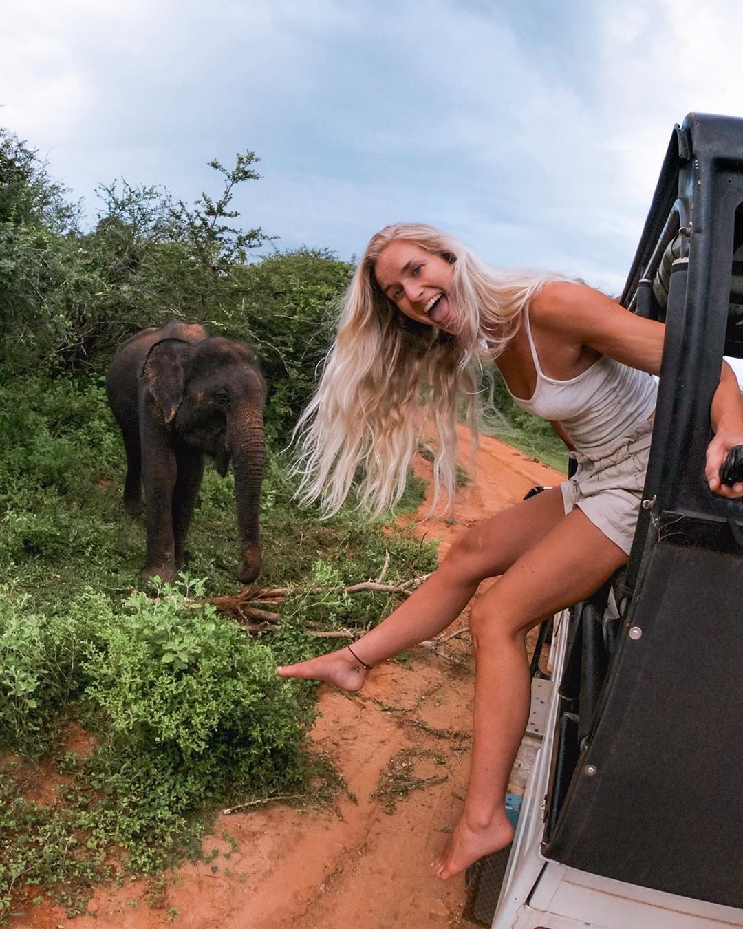 Mujer sonriente asomada desde un vehículo de safari, cerca de un elefante joven en un camino de tierra rodeado de vegetación.