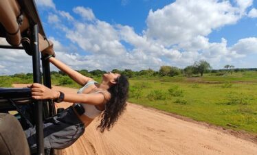Mujer disfrutando de un safari, colgada del vehículo sobre un camino de tierra rodeado de vegetación bajo un cielo azul con nubes.