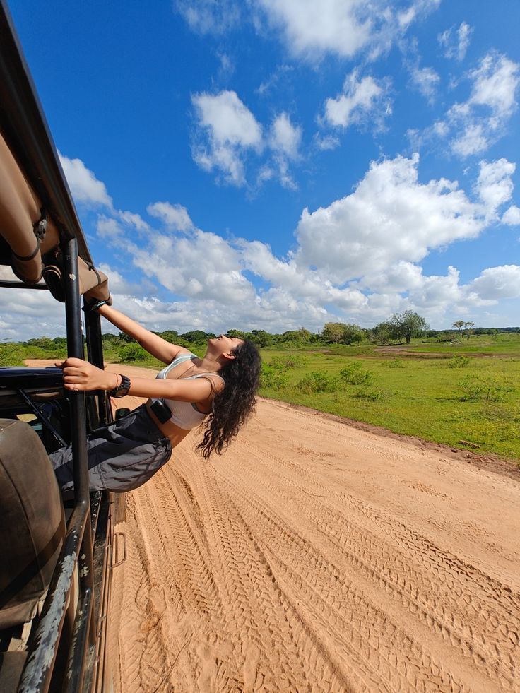 Mujer disfrutando de un safari, colgada del vehículo sobre un camino de tierra rodeado de vegetación bajo un cielo azul con nubes.