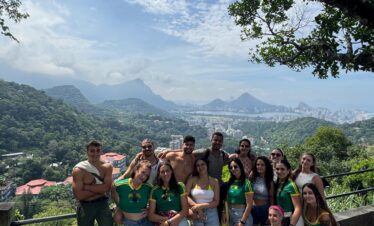 Grupo de jóvenes posando juntos en un mirador con vista panorámica de una ciudad montañosa rodeada de vegetación, bajo un cielo parcialmente nublado.