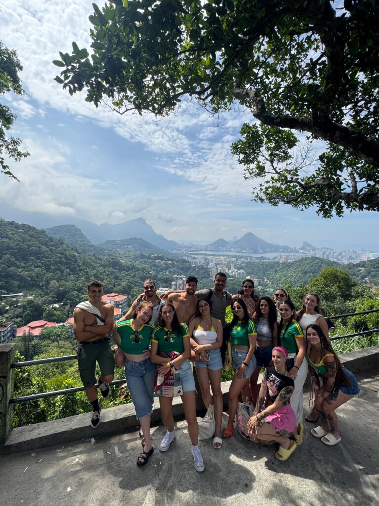 Grupo de jóvenes posando juntos en un mirador con vista panorámica de una ciudad montañosa rodeada de vegetación, bajo un cielo parcialmente nublado.