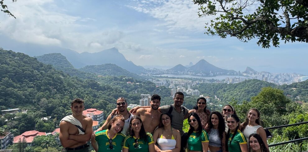Grupo de jóvenes posando juntos en un mirador con vista panorámica de una ciudad montañosa rodeada de vegetación, bajo un cielo parcialmente nublado.