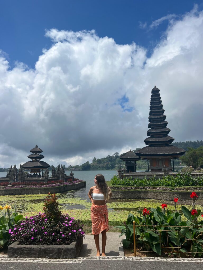 Mujer posando frente al templo Ulun Danu Beratan en Bali, rodeada de flores y estanques bajo un cielo parcialmente nublado.