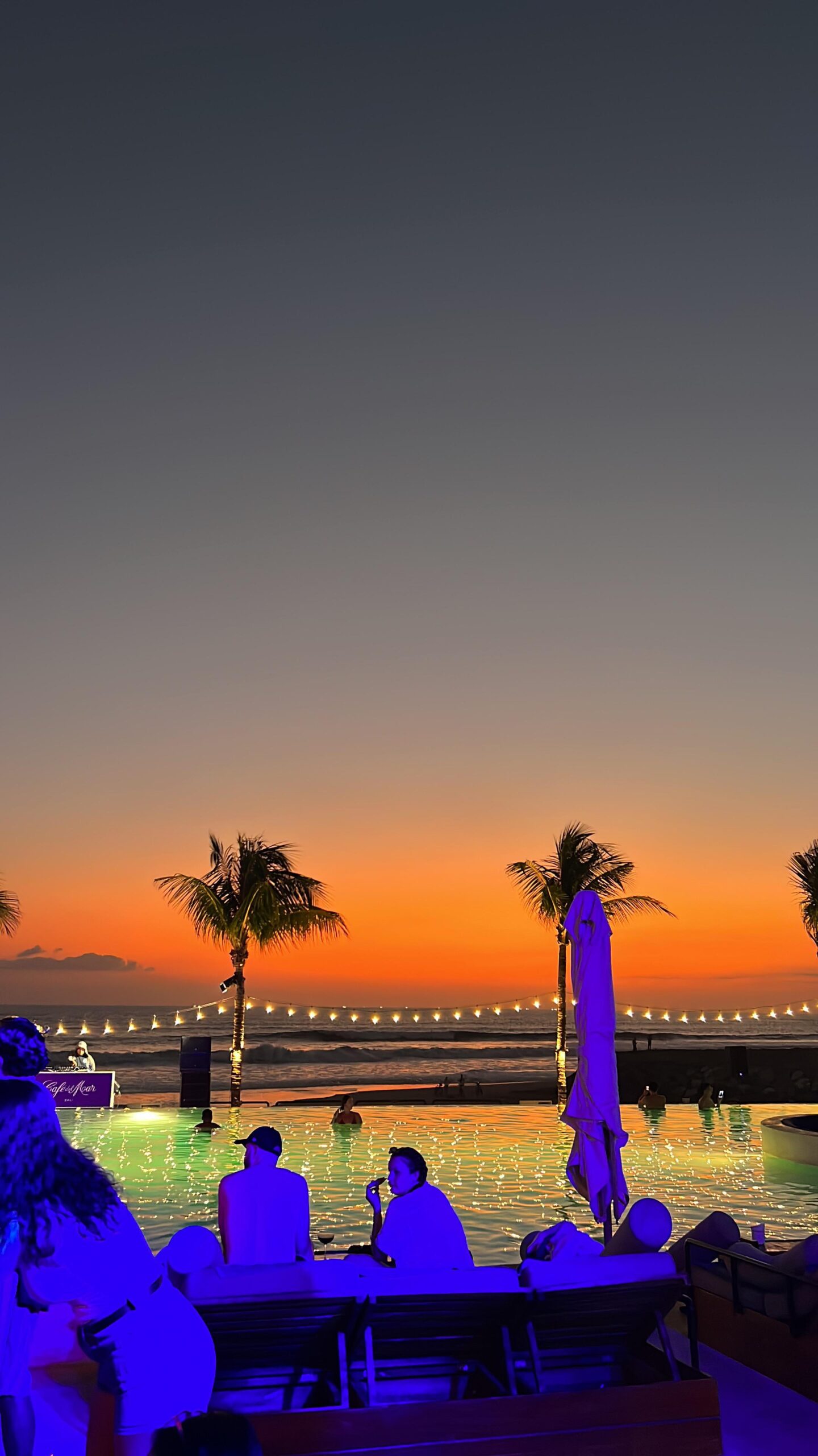 Personas disfrutando de una piscina frente al mar al atardecer, con luces decorativas y palmeras iluminadas.