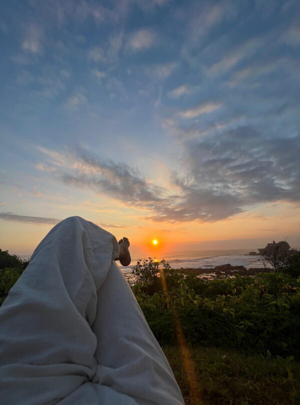 Persona recostada viendo el amanecer o atardecer frente al mar, con vista a un cielo parcialmente nublado y vegetación costera.