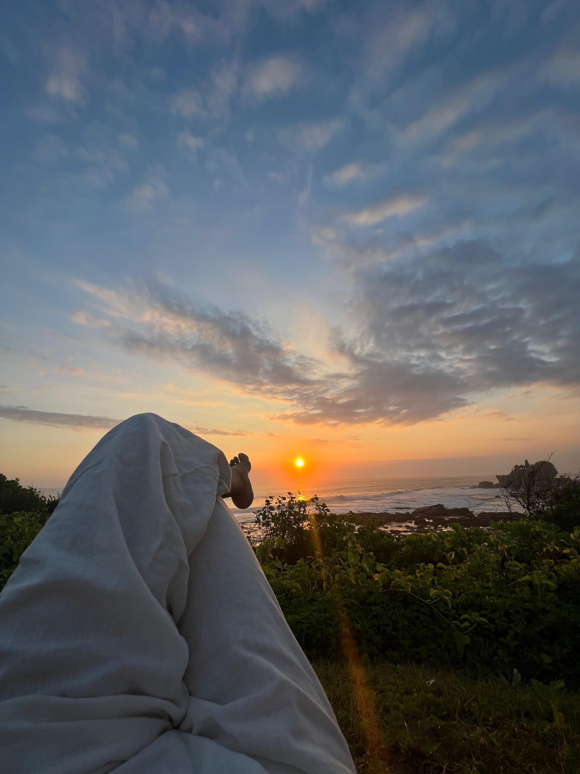 Persona recostada viendo el amanecer o atardecer frente al mar, con vista a un cielo parcialmente nublado y vegetación costera.