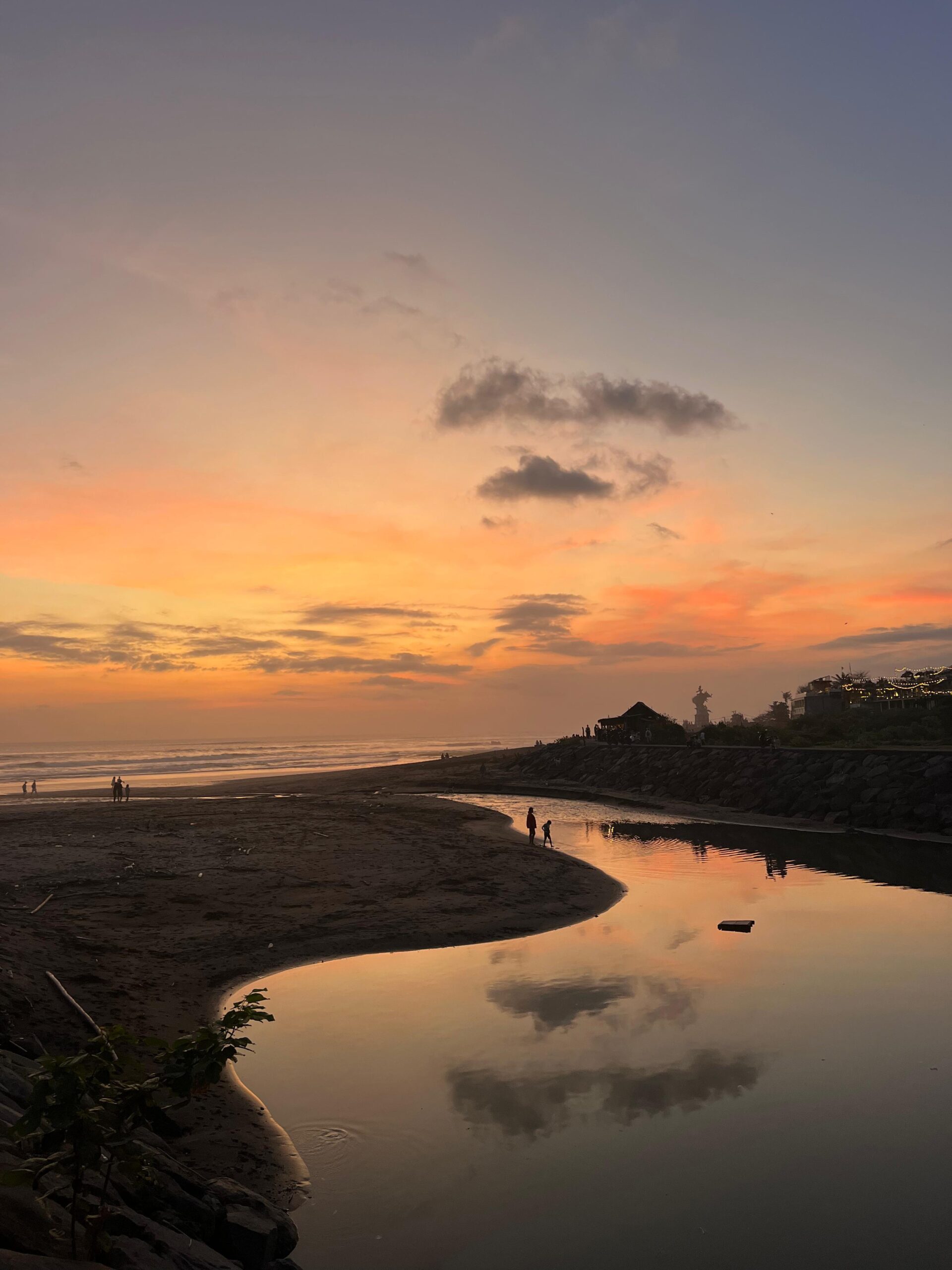 Atardecer en la playa con reflejos en un arroyo, personas caminando y cielo pintado en tonos cálidos.