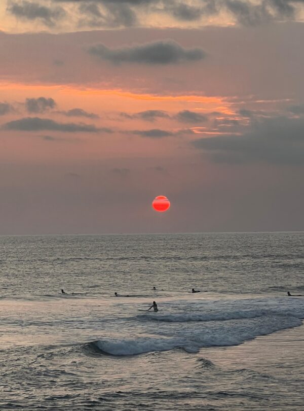 Surfistas en el mar al atardecer con un sol rojo intenso descendiendo sobre el horizonte.