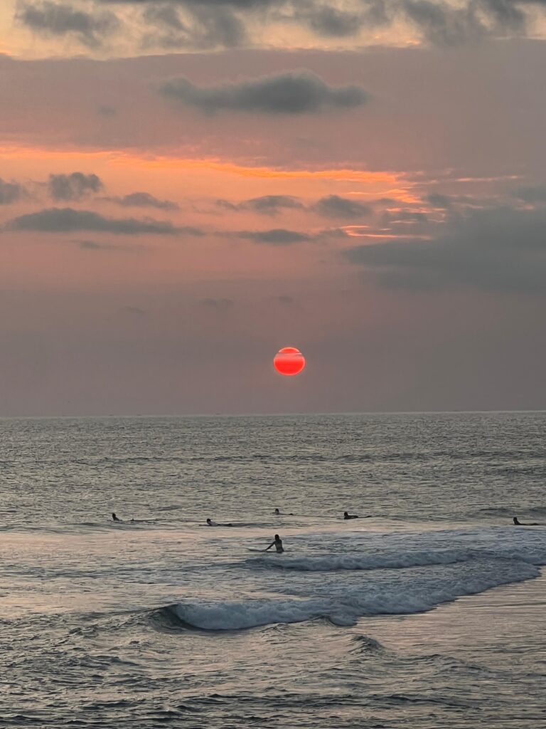 Surfistas en el mar al atardecer con un sol rojo intenso descendiendo sobre el horizonte.