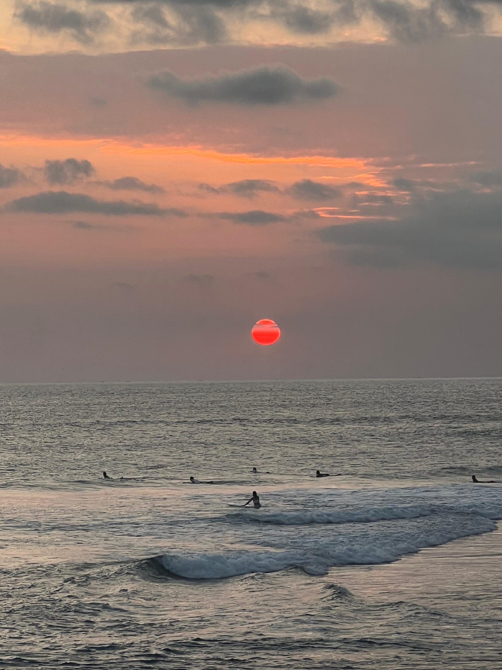 Surfistas en el mar al atardecer con un sol rojo intenso descendiendo sobre el horizonte.