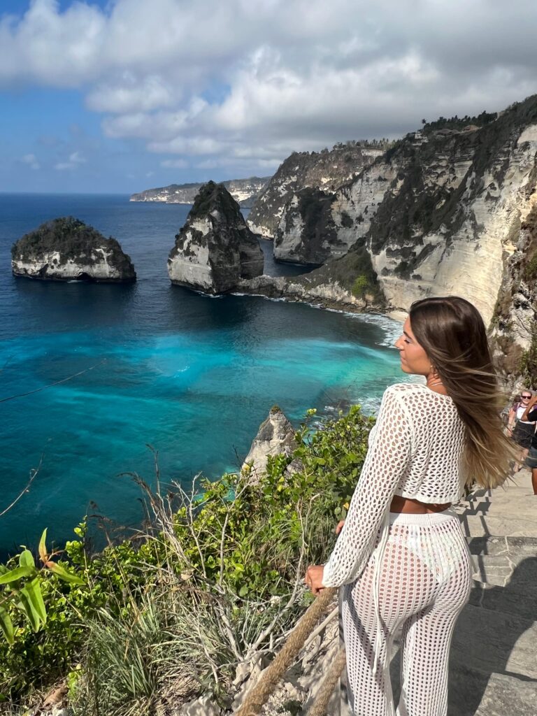 Mujer mirando hacia los acantilados y formaciones rocosas del mar en Nusa Penida, Bali, con aguas turquesas y cielo parcialmente nublado