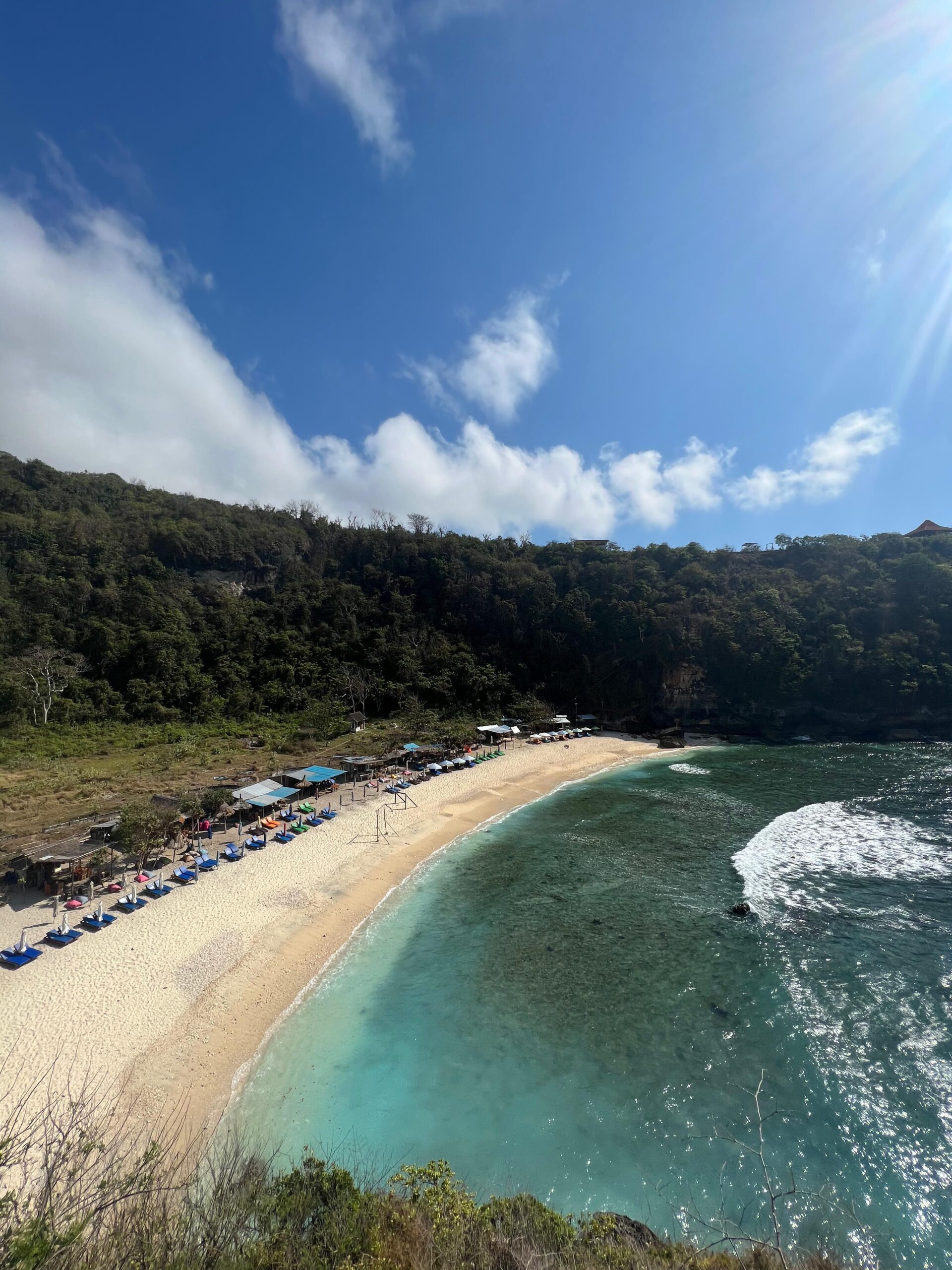 Playa de arena clara con sombrillas y tumbonas alineadas frente a un mar turquesa, rodeada de vegetación y acantilados.