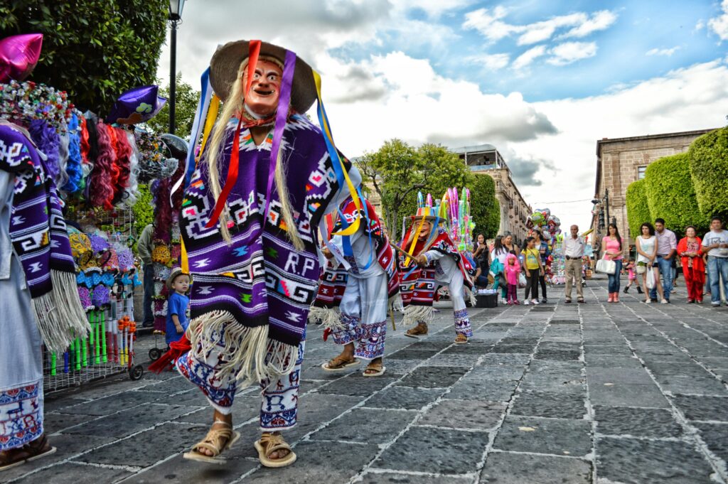 Danzantes tradicionales con máscaras y vestimenta colorida participan en una celebración cultural en una plaza mexicana.