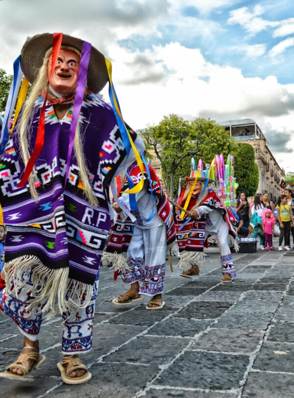 Danzantes tradicionales con máscaras y vestimenta colorida participan en una celebración cultural en una plaza mexicana.