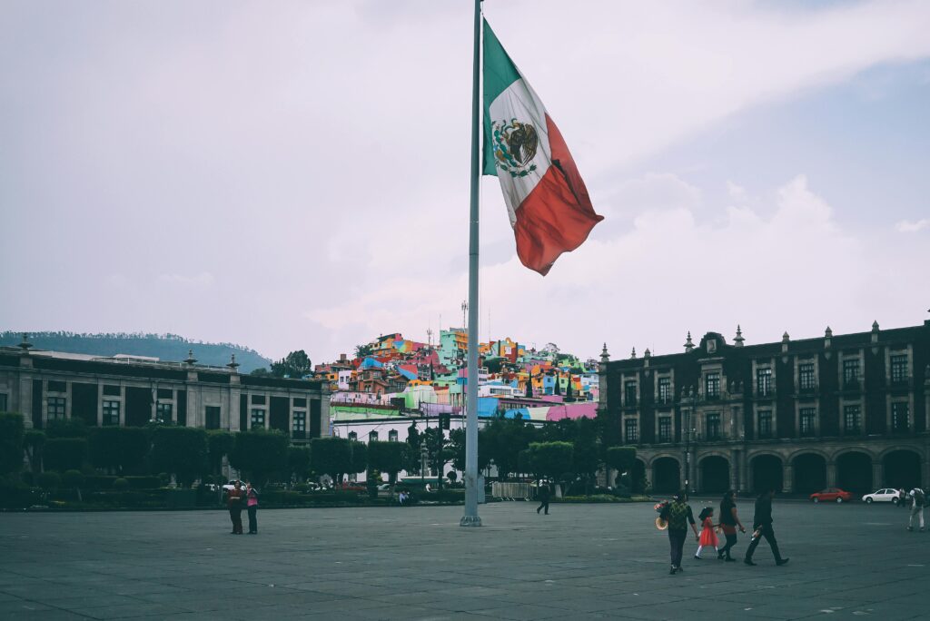 Plaza con bandera de México ondeando, edificios coloniales y casas coloridas en una colina al fondo.