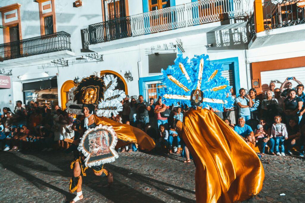 Danzantes con trajes tradicionales y penachos coloridos participan en una festividad nocturna en una calle empedrada frente al público.