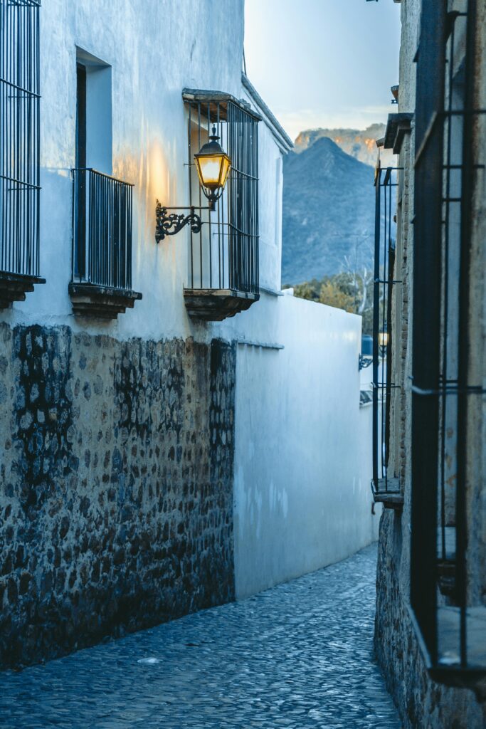 Callejón empedrado con faroles encendidos y casas coloniales con montañas al fondo.